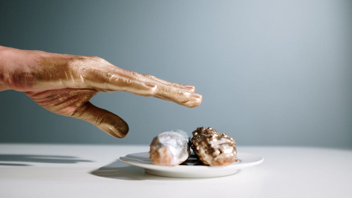 A golden-painted hand reaches towards artfully decorated pastries on a white plate.